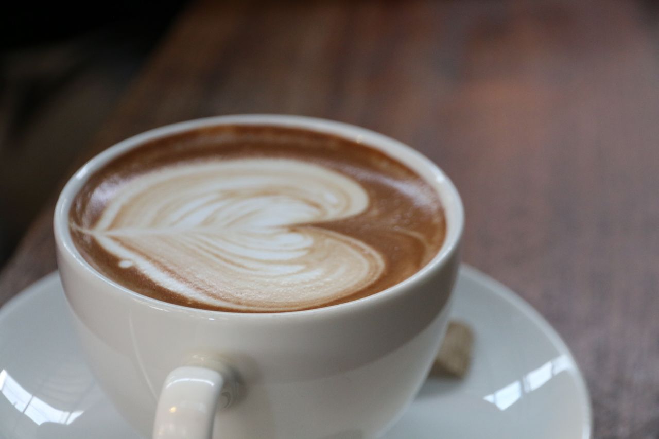 gallery-01 Close-up of a latte with heart-shaped art in a white cup on a wooden table.