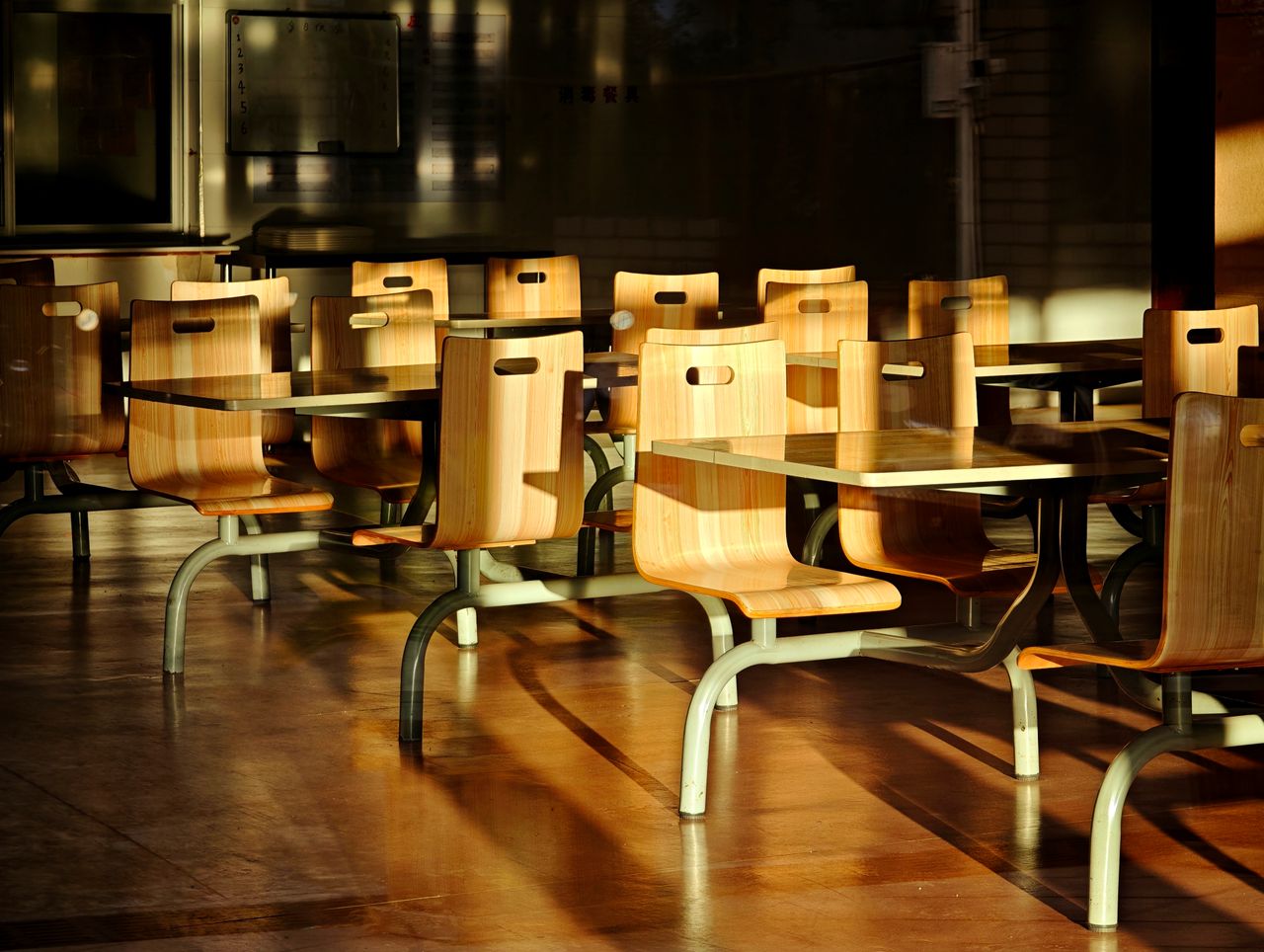 gallery-03 Empty wooden chairs and tables in a sunlit cafeteria setting, casting dramatic shadows.