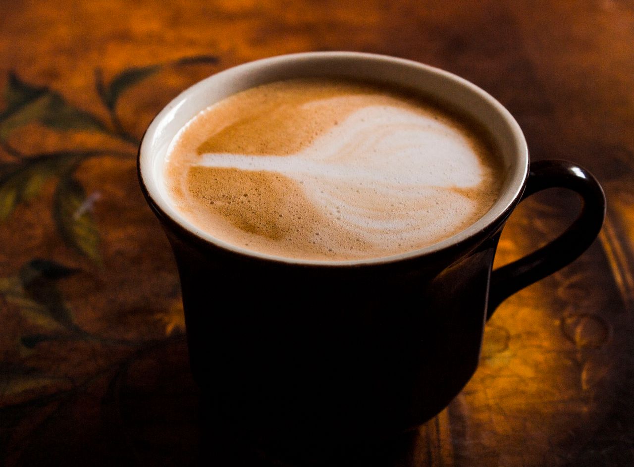 our-story A close-up of a cappuccino in a black mug with latte art, against a warm, artistic background.